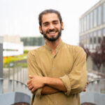 Happy joyful hipster guy posing for camera outside. Young man in casual with bun and stubble standing in urban settings, looking at camera and smiling. Male portrait concept